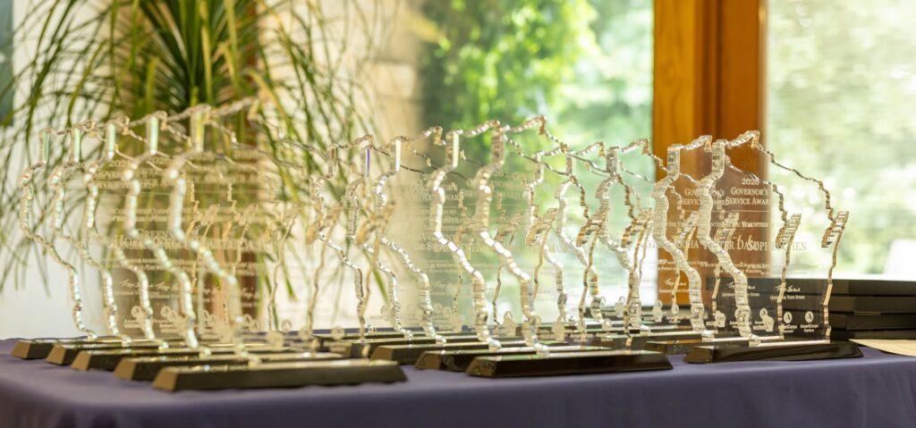 Photo of Governor's Service Awards sitting on a table to be handed out. The award is etched glass in the shape of wisconsin, standing up perpendicular from the base.
